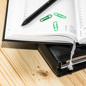 Open planner with black pen and green paperclips on a wooden desk, with a black leather-bound cover beneath.