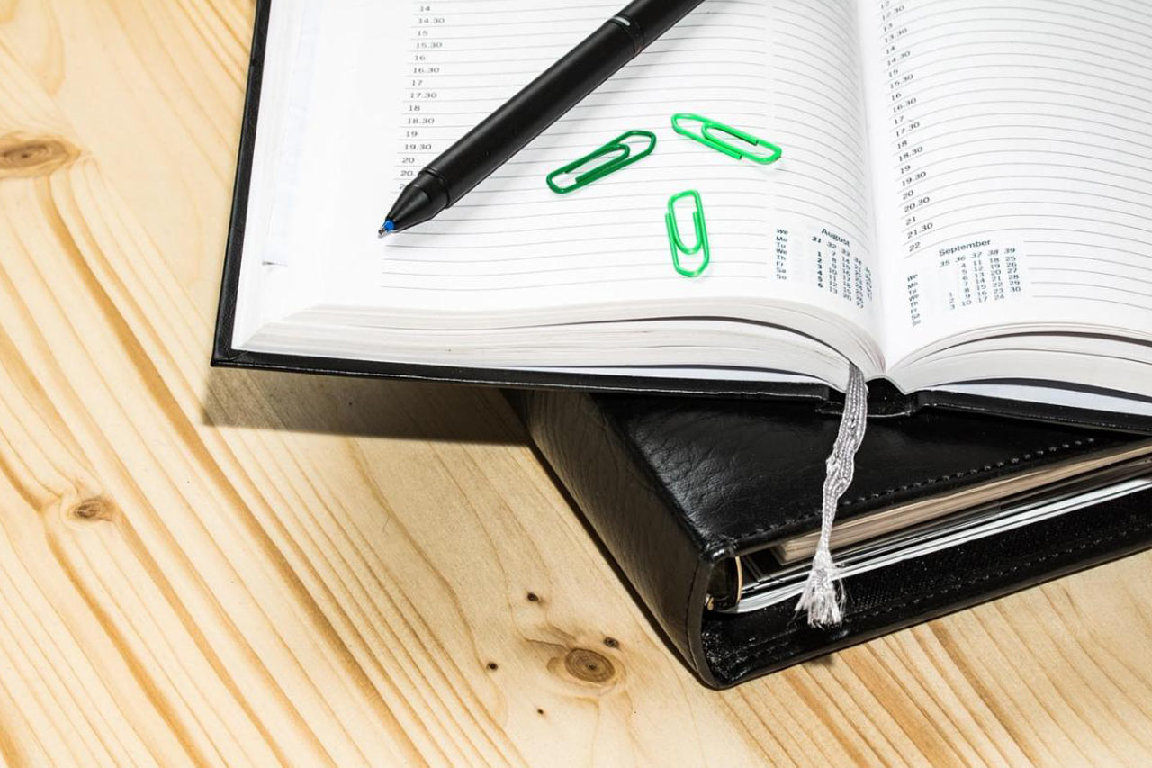 Open planner with black pen and green paperclips on a wooden desk, with a black leather-bound cover beneath.