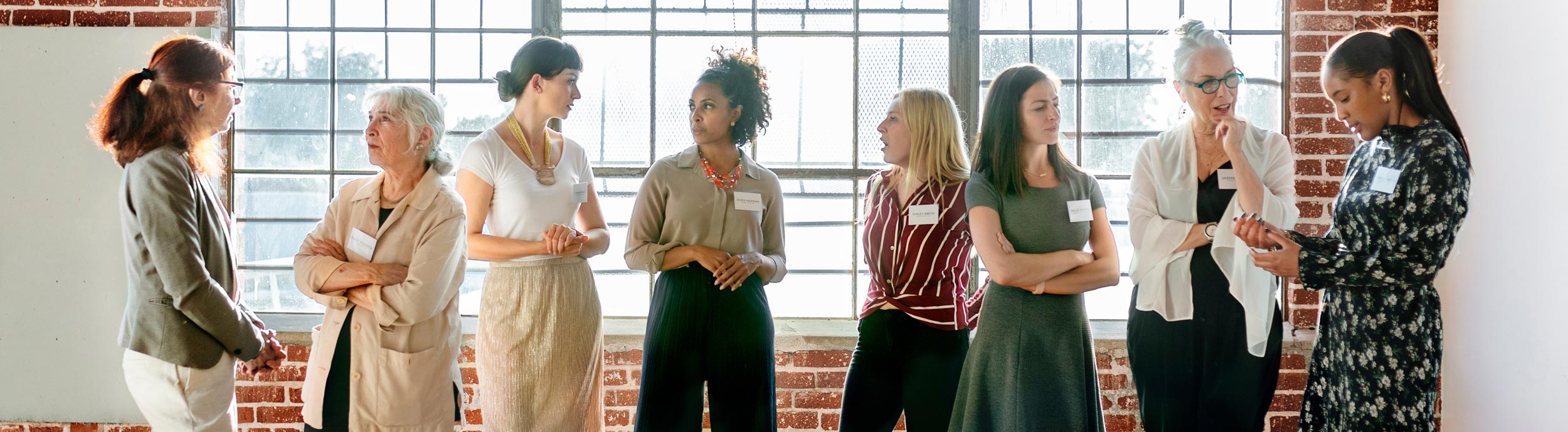 A group of diverse women standing and conversing near a large window in an industrial-style room, each wearing a name tag.